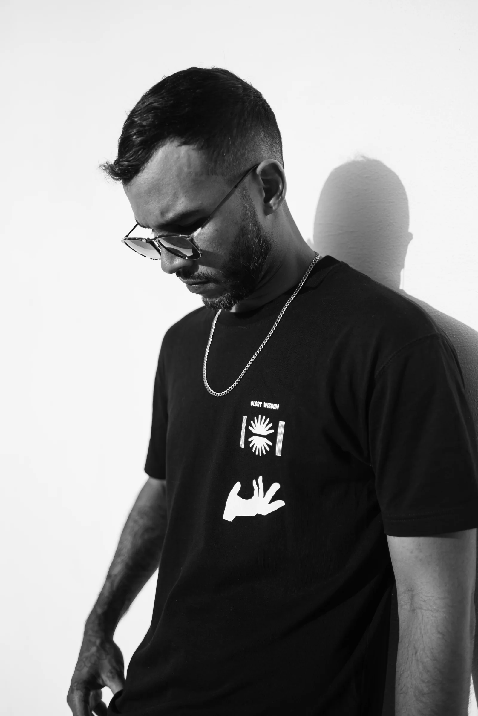 Black-and-white portrait of Carlos wearing sunglasses, looking down beside a sunlit wall.